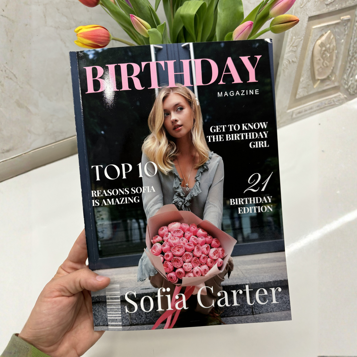 A hand holds a copy of Sofia Carter's 'Birthday Magazine' featuring a cover with pink roses and text about Sofia Carter. Tulips are visible at the top of the frame against a tiled wall background.