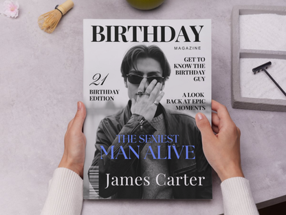 A person holds a customized 'Birthday Magazine' featuring a cover photo of James Carter with the headline 'The Sexiest Man Alive'. The scene is styled as a flat-lay on a light gray textured surface with a whisk, a green object, and a small box in the background.