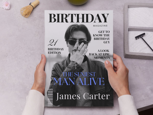 A person holds a customized 'Birthday Magazine' featuring a cover photo of James Carter with the headline 'The Sexiest Man Alive'. The scene is styled as a flat-lay on a light gray textured surface with a whisk, a green object, and a small box in the background.