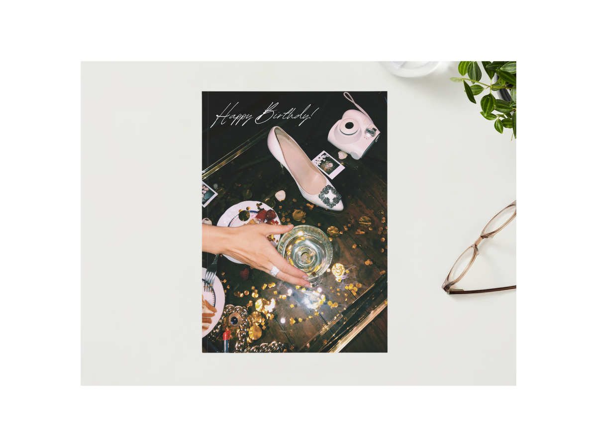 A flat-lay styled product photo of a square greeting card on a light gray surface. The card features a dark, artistic flat-lay design with a hand holding a glass, a white pointed shoe, a camera, and gold glitter, with 'Happy Birthday!' written at the top.