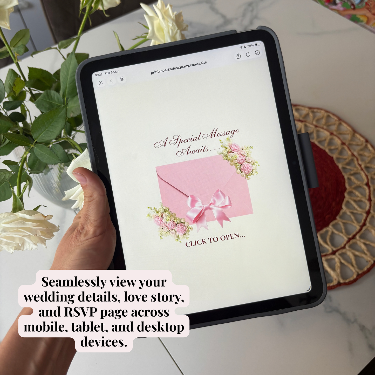 A hand holds a tablet showing a wedding invitation design with a pink envelope and flowers. The scene includes white roses, a marble surface, and a woven placemat.