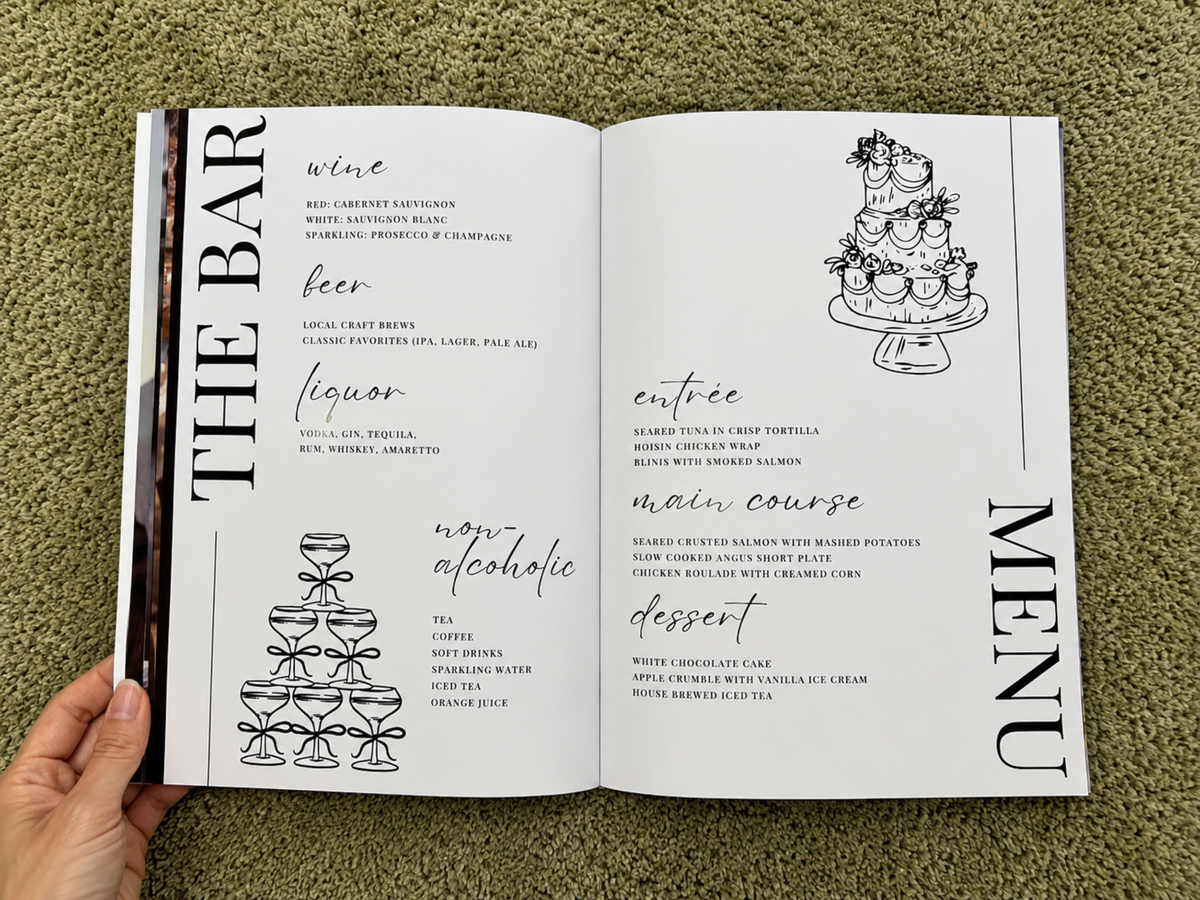An open printed menu book or recipe booklet laid on green grass, held at the bottom by a person's hand. The pages show black text and line drawings of a champagne tower, wine, beer, liquor, entree, main course, and dessert under the heading THE BAR MENU.