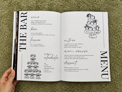 An open printed menu book or recipe booklet laid on green grass, held at the bottom by a person's hand. The pages show black text and line drawings of a champagne tower, wine, beer, liquor, entree, main course, and dessert under the heading THE BAR MENU.